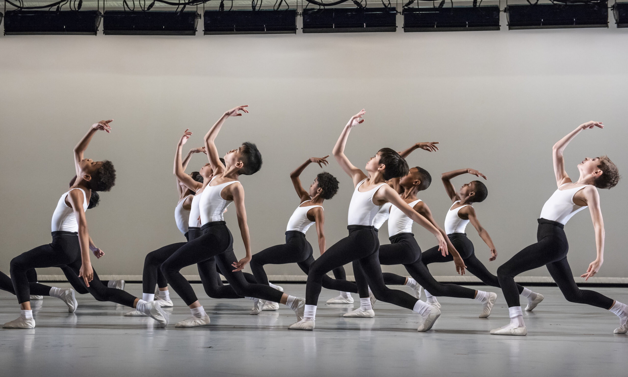 A group of dancers, dressed in white shirts and black leggings, perform synchronized movements on stage. They look upward, striking dynamic poses with one arm extended and one leg bent, emphasizing precision and grace, against a plain background.